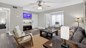 A modern living room with a ceiling fan, a flat-screen TV mounted on the wall, a sofa, a coffee table, and a lamp. - Serene Mins 2 Sand Beach Brookdale Mayo Clinic Everbank (Jacksonville)
