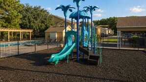 A playground with a slide and climbing structures, surrounded by a fence and a pool. - Serene Mins 2 Sand Beach Brookdale Mayo Clinic Everbank (Jacksonville)