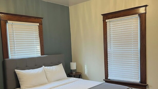 Ein Schlafzimmer mit einem Bett, zwei Fenstern mit Jalousien, einem Nachttisch mit Lampe und einer Deckenleuchte.