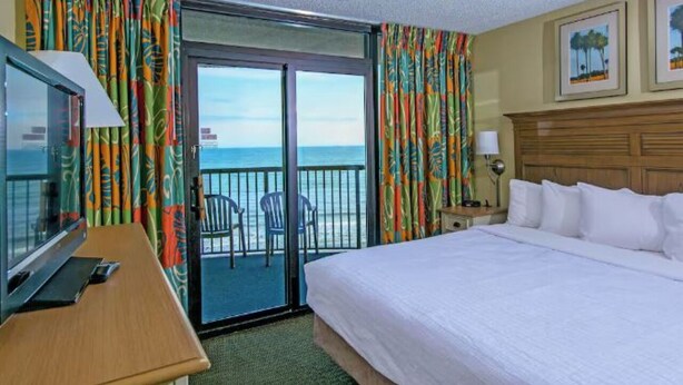 Habitación de hotel con cama, escritorio con ordenador, televisor y balcón con sillas.