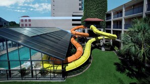 Zona de piscina con tobogán, área de descanso cubierta y vista a un edificio alto con techo verde.