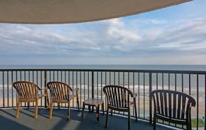 Un balcón con cuatro sillas de madera y una mesa pequeña, con vista a una playa y una vista despejada del océano.
