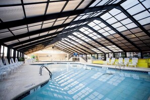 Piscina interior com teto de vidro, cadeiras de descanso brancas e vista clara para a área da piscina.