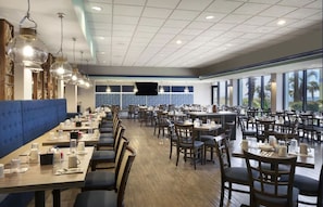 Ein geräumiges Restaurant mit mehreren Tischen und Stühlen, einer blauen gepolsterten Bank und großen Fenstern mit Blick auf Palmen.