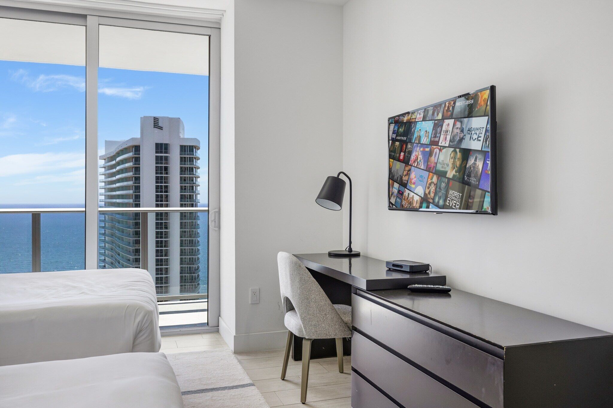 Habitación de hotel con balcón, escritorio, silla y televisor de pared mostrando carteles de películas.
