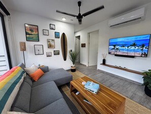 Ein modernes Wohnzimmer mit einem grauen Sofa, einem Couchtisch mit Büchern, einem an der Wand befestigten Surfbrett und einem Fernseher, der eine Strandansicht zeigt.