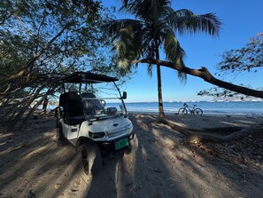 Un carrito de golf estacionado en una playa arenosa con vista al océano y veleros en la distancia.