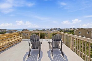 A deck with two chairs facing a beach view, wooden railings, and houses in the background.