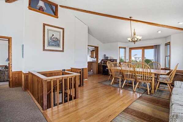 A spacious dining area with a wooden table and chairs, a chandelier, and a view of the outdoors through large windows.