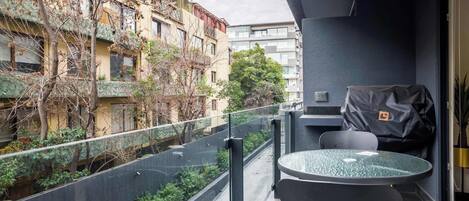 A balcony with a glass table and chairs, a grill, and a view of a building with trees.