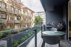 Ein Balkon mit Glastisch und Stühlen, einem Grill und Blick auf ein Gebäude mit Bäumen.