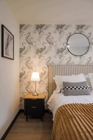 Ein Schlafzimmer mit einem Bett, einem Nachttisch, einer Lampe und einer Wand mit ornamentalem Wandpapier.