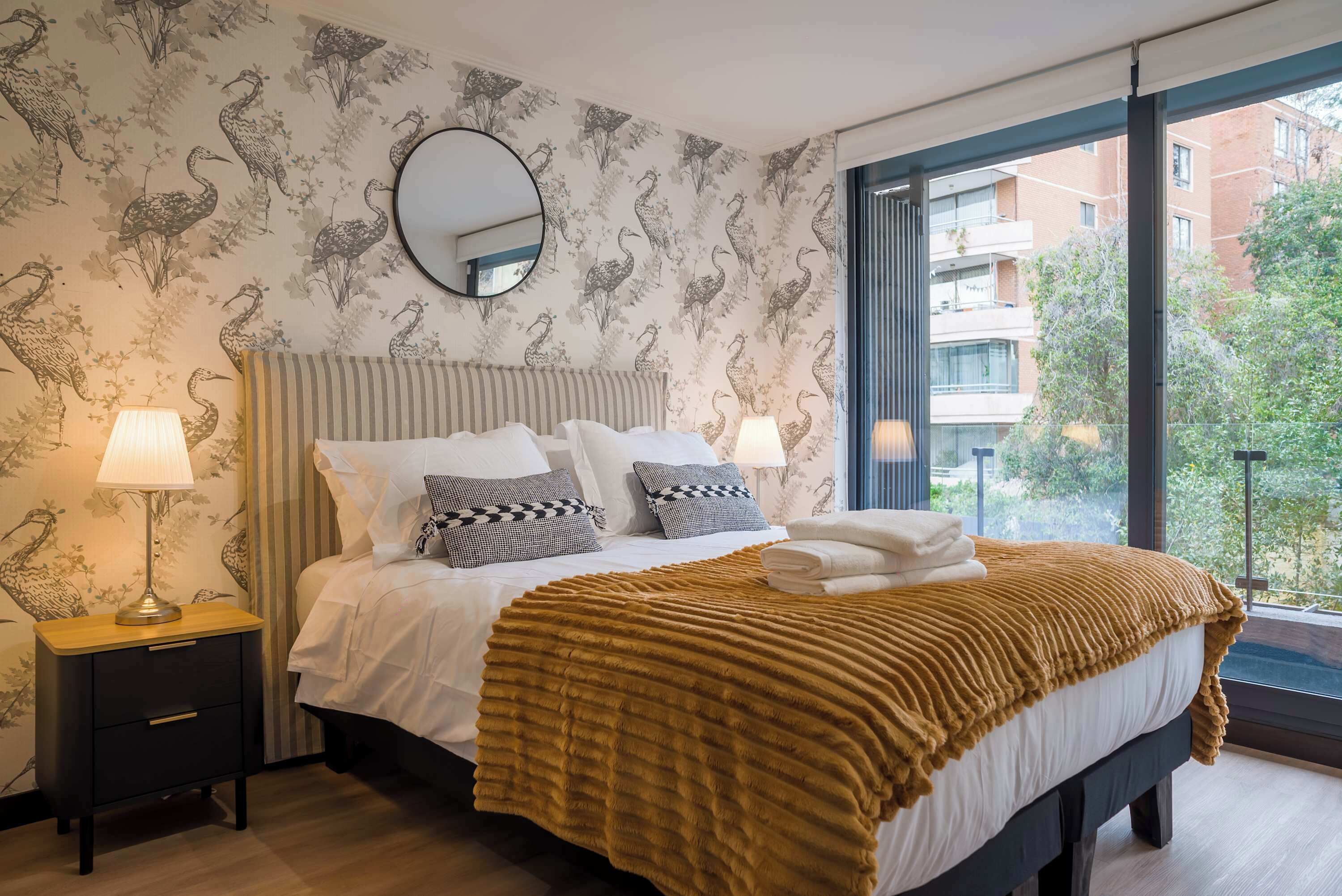 A hotel room with a large bed, two bedside lamps, a mirror above the bed, and a window with a view of buildings and greenery.