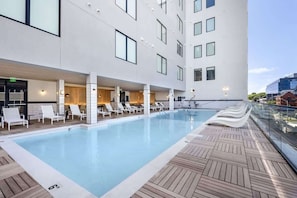 Una piscina en la azotea con sillones reclinables, una terraza de madera y un edificio moderno con grandes ventanales.