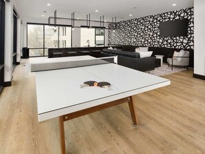 Una mesa de ping pong en un espacio habitable moderno con una cocina al fondo, un sofá y un televisor en la pared.