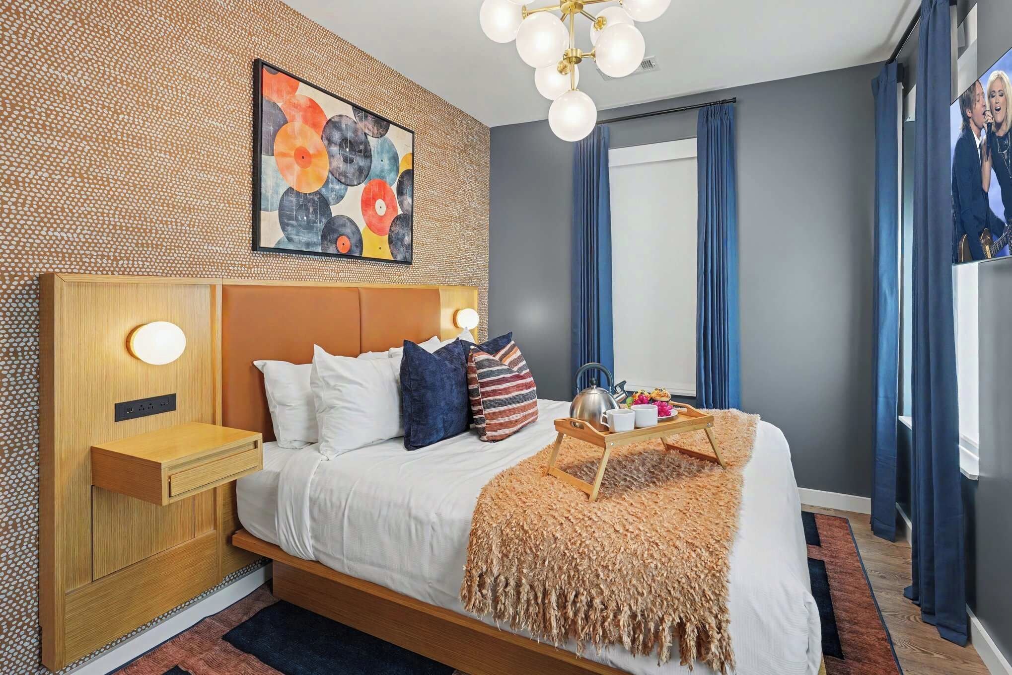 A modern bedroom with a wooden headboard, a bed with white bedding, a decorative wall art piece, a bedside table with a lamp, and a window with curtains.