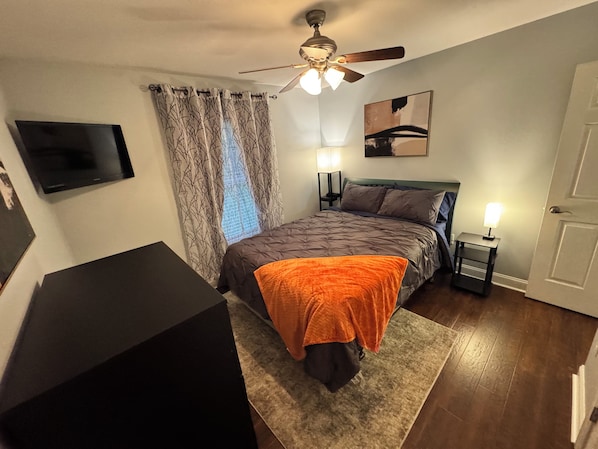 A bedroom with a bed, a ceiling fan, a flat-screen TV, a nightstand with a lamp, and a painting on the wall.
