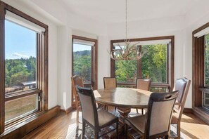 A dining area with a round table and chairs, large windows with a view of greenery, and a chandelier. - Chamonix 50 | Ski-in/out 2BR Condo with Mountain Views, Pool, Hot Tub, and Gym (Snowmass Village)