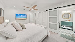 A bedroom with a bed, a ceiling fan, a wall-mounted TV, a bathroom with a vanity, and a sliding door closet. - New! Blue Haven - Heated Saltwater Pool/Spa, 1 Mile to the Beach! (Naples)