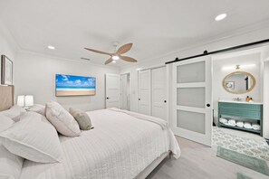 Ein Schlafzimmer mit Bett, Deckenventilator, Wand-TV, Badezimmer mit Waschtisch und Schiebetürenschrank.