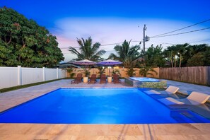 A swimming pool with lounge chairs and umbrellas, surrounded by a wooden fence and palm trees. - New! Blue Haven - Heated Saltwater Pool/Spa, 1 Mile to the Beach! (Naples)