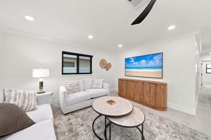 A modern living room with a white sofa, a round coffee table, a wooden cabinet, and a large beach-themed painting on the wall. - New! Blue Haven - Heated Saltwater Pool/Spa, 1 Mile to the Beach! (Naples)