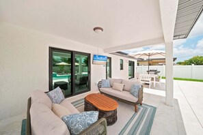 A covered outdoor seating area with a pool view, a sofa, a coffee table, and a TV screen. - New! Blue Haven - Heated Saltwater Pool/Spa, 1 Mile to the Beach! (Naples)
