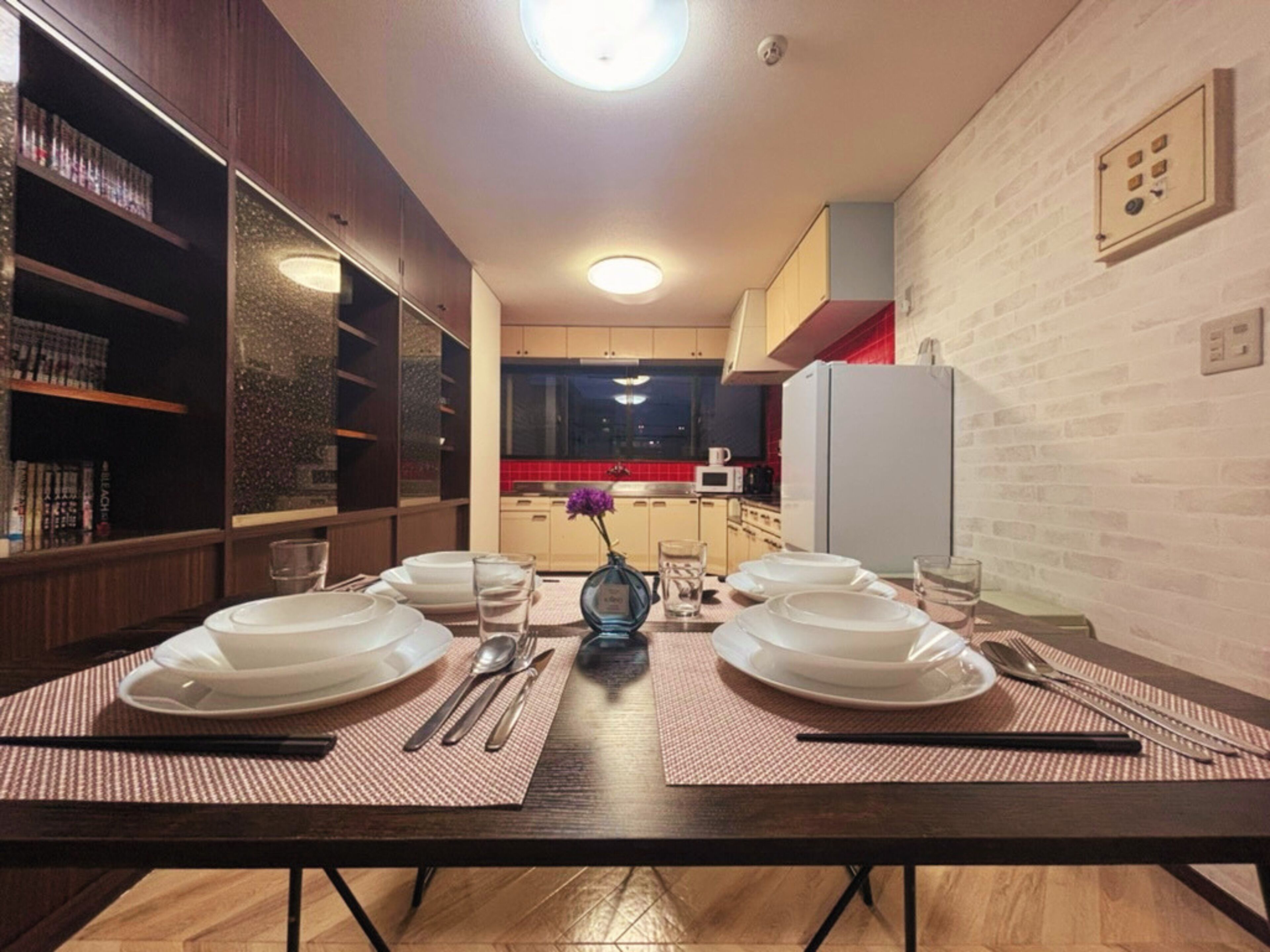 A dining area with a long table set for four, surrounded by bookshelves and a kitchen in the background.