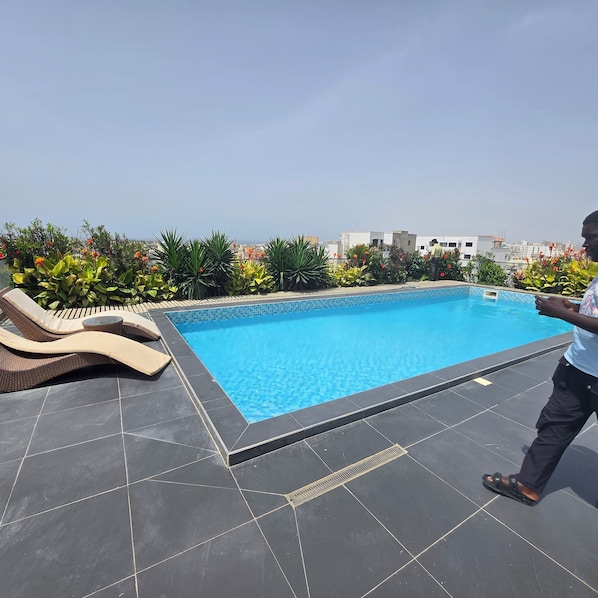 Una piscina en la azotea con vistas despejadas del paisaje urbano, rodeada de abundante vegetación y un área de descanso con una sola silla.