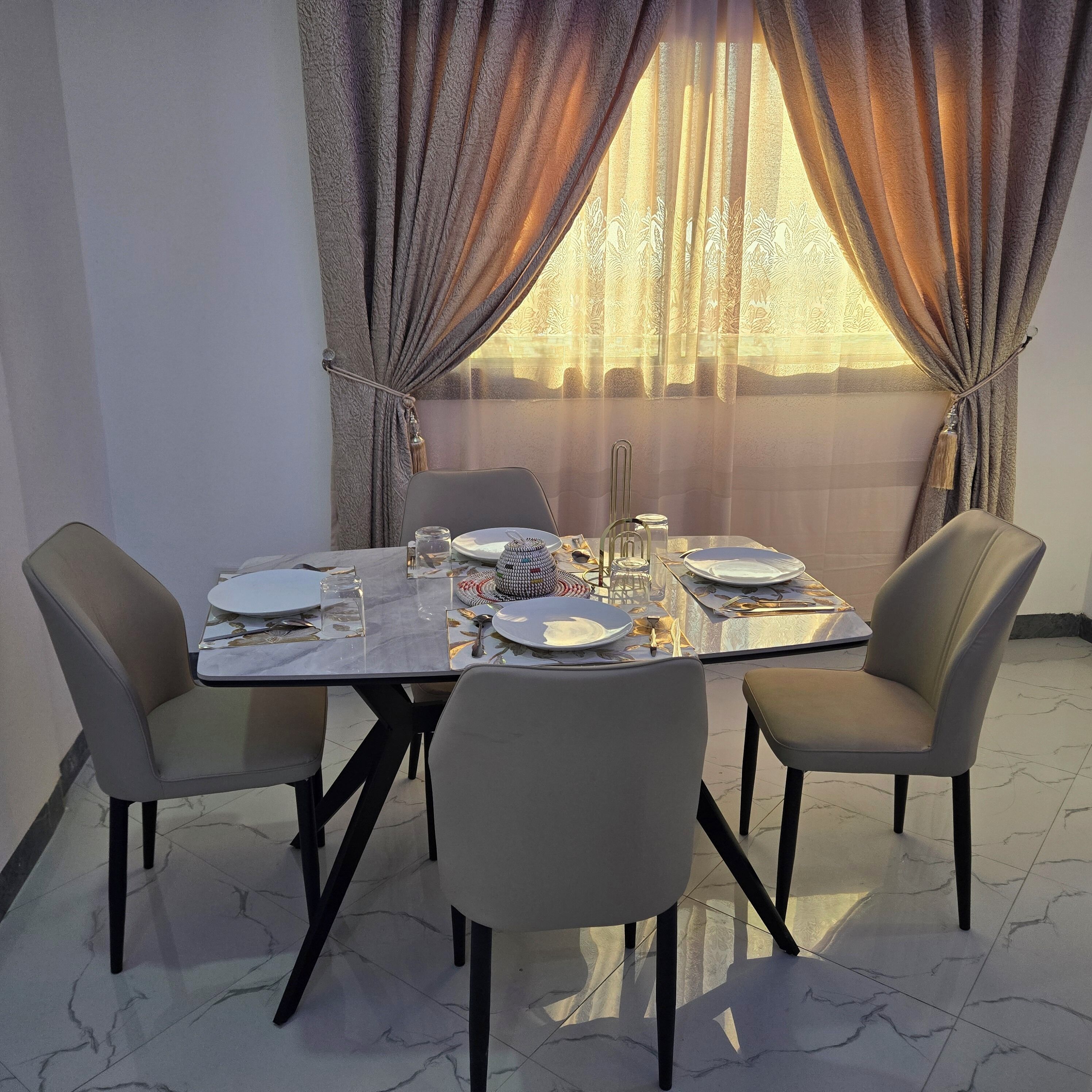Une table à manger pour quatre personnes, avec des chaises, une fenêtre avec des rideaux et un sol en marbre.