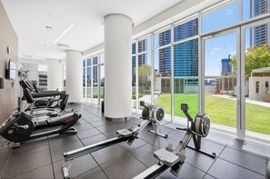 Ein Fitnessraum mit Herz-Kreislauf-Geräten, großen Fenstern und Blick auf Gebäude und Grünflächen.