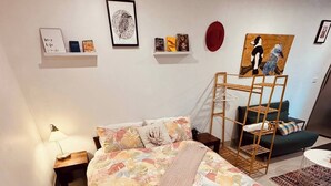 Un dormitorio con cama, mesita de noche, lámpara, un cuadro enmarcado, un sombrero rojo, una escalera de madera y un sofá verde.
