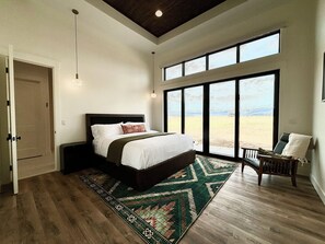 Un dormitorio con una cama grande, vistas al exterior a través de una puerta corrediza, piso de madera y una silla junto a la ventana.