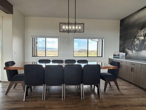 A dining area with a wooden table and six chairs, large windows with a view of the outdoors, and a modern pendant light. - RollingTetonRetreat_Hot tub_Sauna_Patio_FamilyFriendly (Tetonia)
