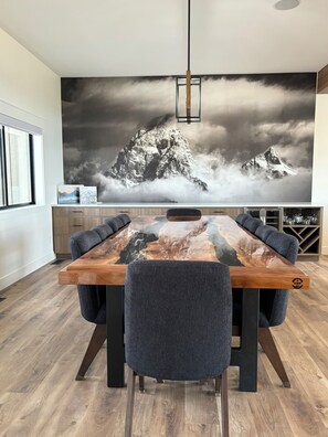 A modern dining area with a long wooden table, six upholstered chairs, and a large mountain landscape mural on the wall. - RollingTetonRetreat_Hot tub_Sauna_Patio_FamilyFriendly (Tetonia)