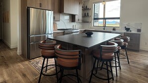 A modern kitchen with wooden cabinets, a stainless steel refrigerator, a central island with bar stools, and a large window offering a view. - RollingTetonRetreat_Hot tub_Sauna_Patio_FamilyFriendly (Tetonia)