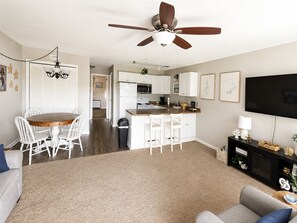A modern living room with a large TV, a ceiling fan, a dining area with a wooden table, and a kitchen with white cabinets. - Surfside Beach Retreat , Pool, Hot Tub, Pickleball & Minutes to the Beach (Surfside Beach)