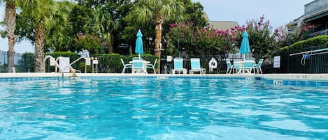 A swimming pool with lounge chairs and umbrellas, surrounded by palm trees and flowering bushes.