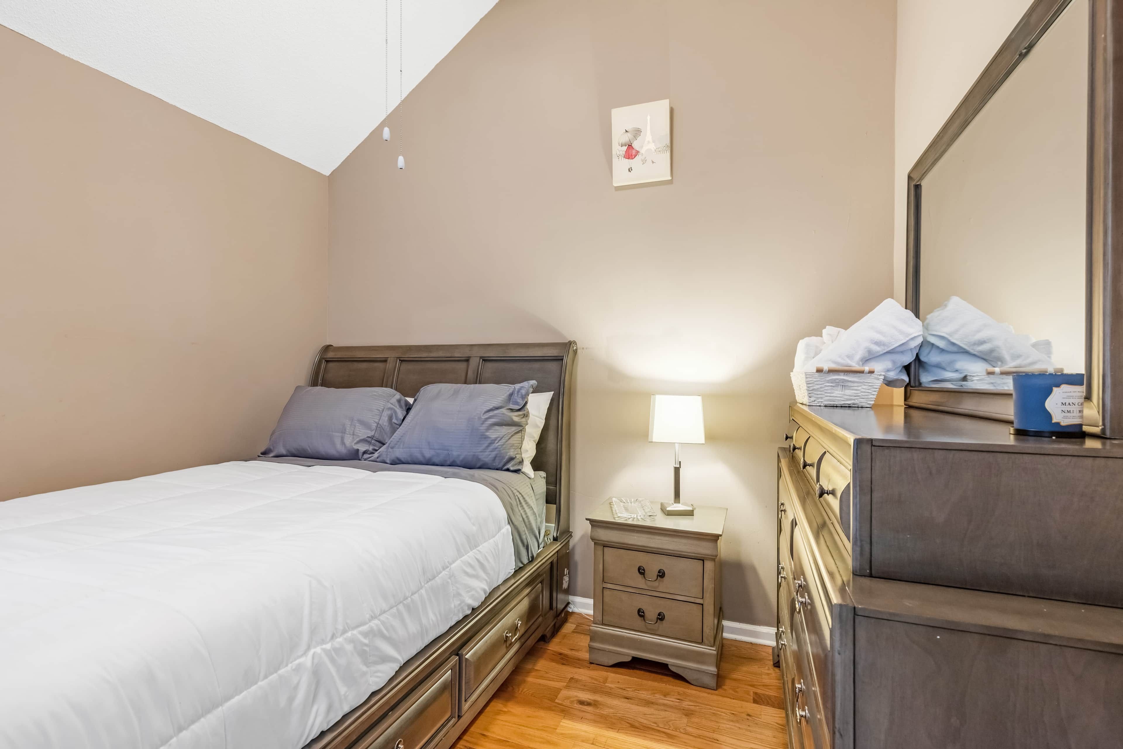 Een slaapkamer met een bed, nachtkastje, lamp, kaptafel en een lijst met een afbeelding aan de muur.