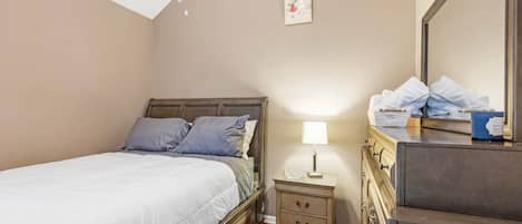 Een slaapkamer met een bed, nachtkastje, lamp, kaptafel en een lijst met een afbeelding aan de muur.