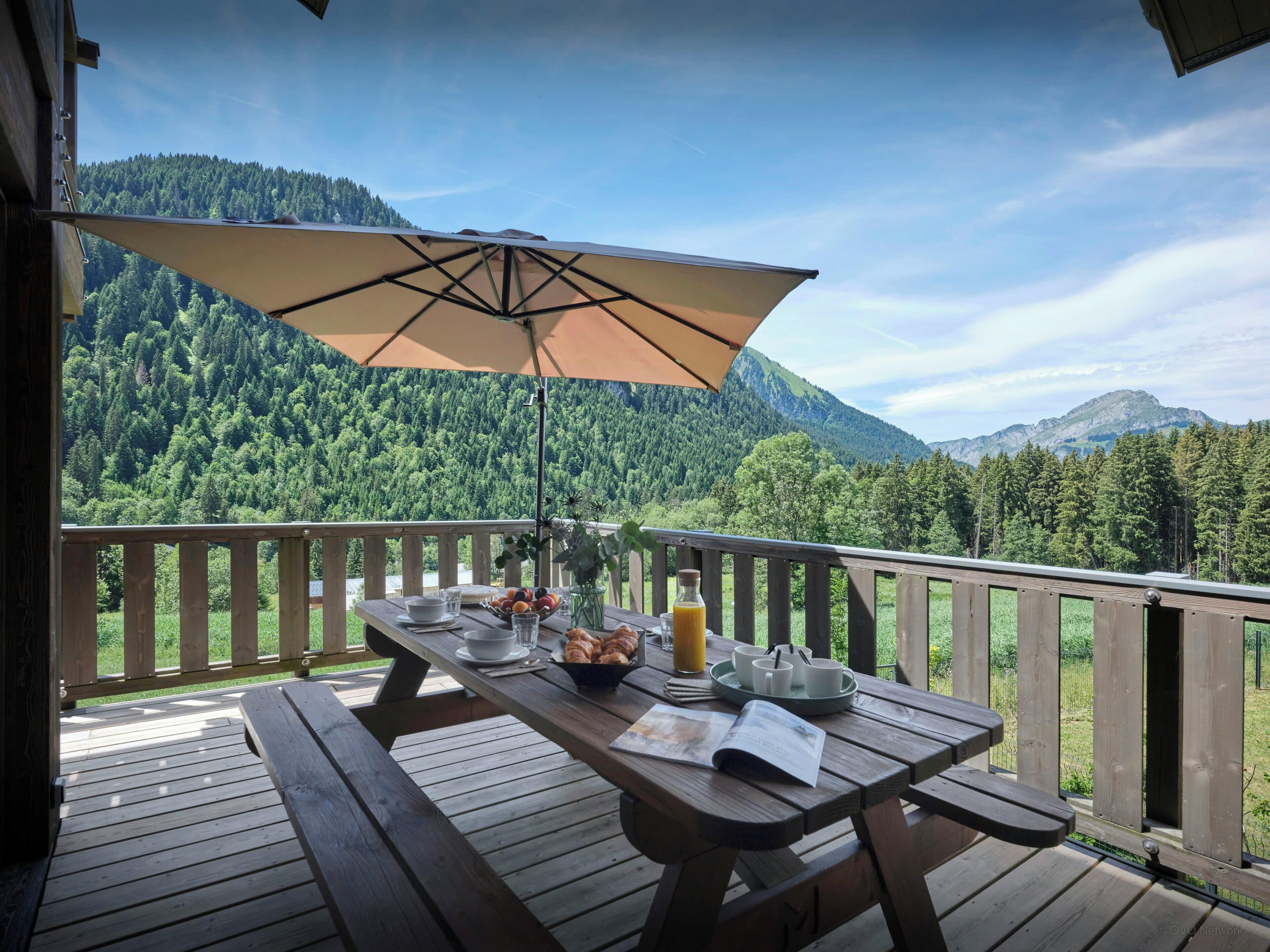 Une terrasse en bois avec une table dressée pour le petit-déjeuner, offrant une vue sur une chaîne de montagnes boisée.
