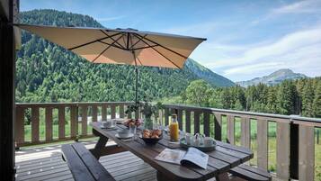 Une terrasse en bois avec une table dressée pour le petit-déjeuner, offrant une vue sur une chaîne de montagnes boisée.