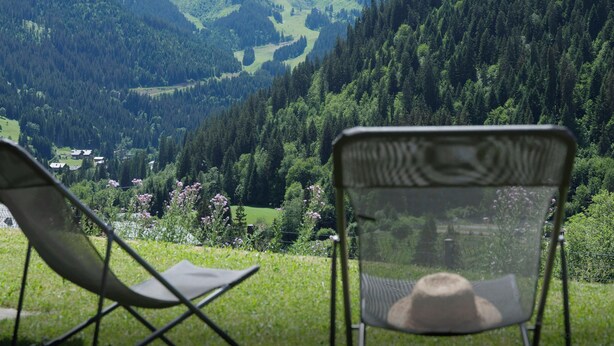 Une chaise sur laquelle est posé un chapeau, d’où l’on aperçoit une vallée avec des montagnes et une maison au loin.