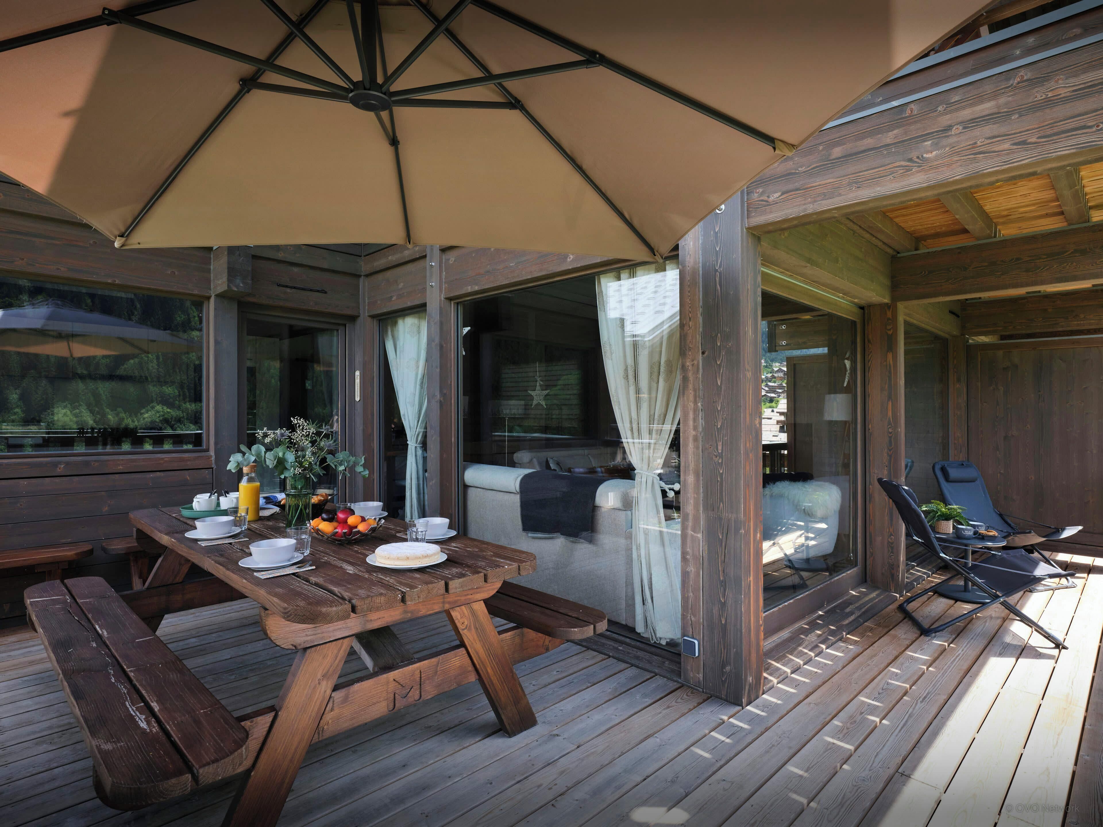 Une terrasse en bois avec une table dressée pour le petit-déjeuner, comprenant des assiettes, des tasses et un panier de fruits.