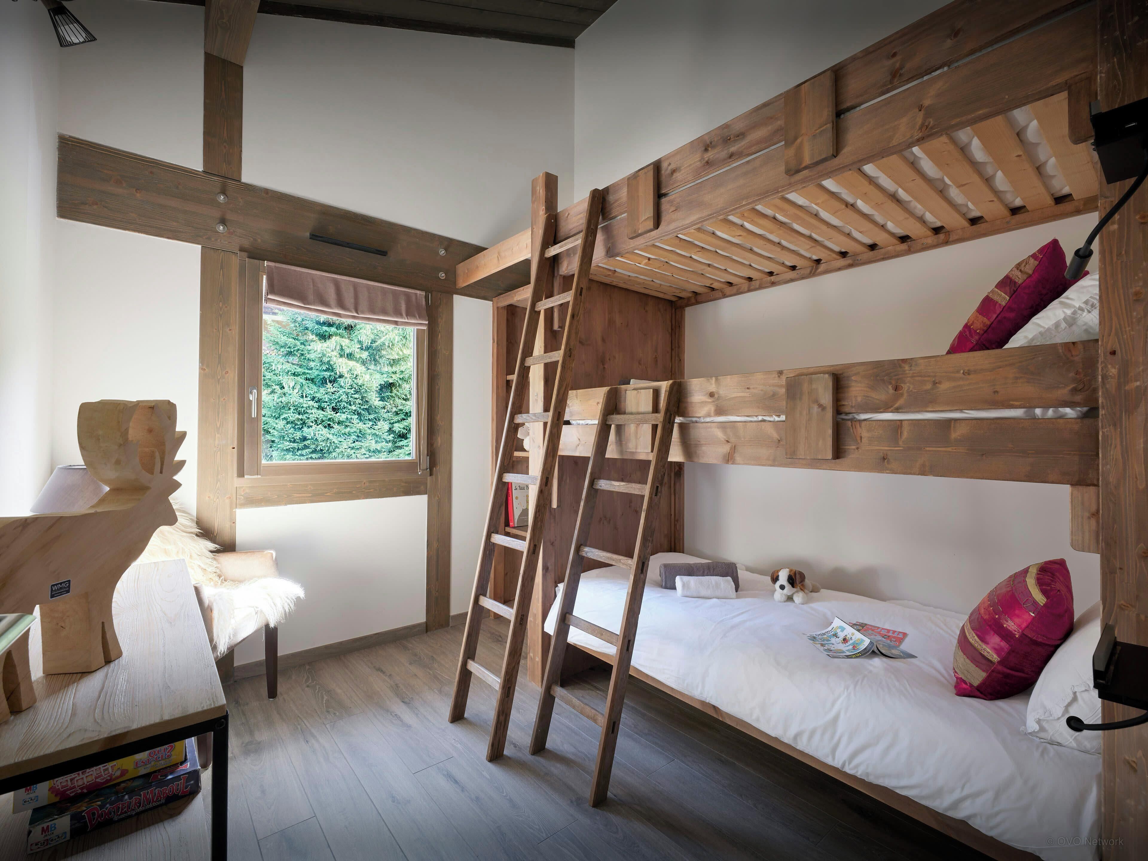 Ein Zimmer mit einem Etagenbett, einer Klapptreppe, einem Schreibtisch mit Stuhl, einem Fenster mit Blick auf Bäume und einem Bücherregal.