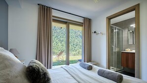 Une chambre avec un grand lit, une vue sur une forêt par une porte coulissante et une salle de bain avec une douche en verre.