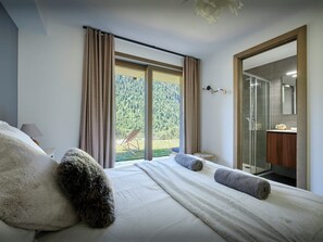 Un dormitorio con una cama grande, vista a un bosque a través de un ventanal corredera y un baño con ducha de vidrio.