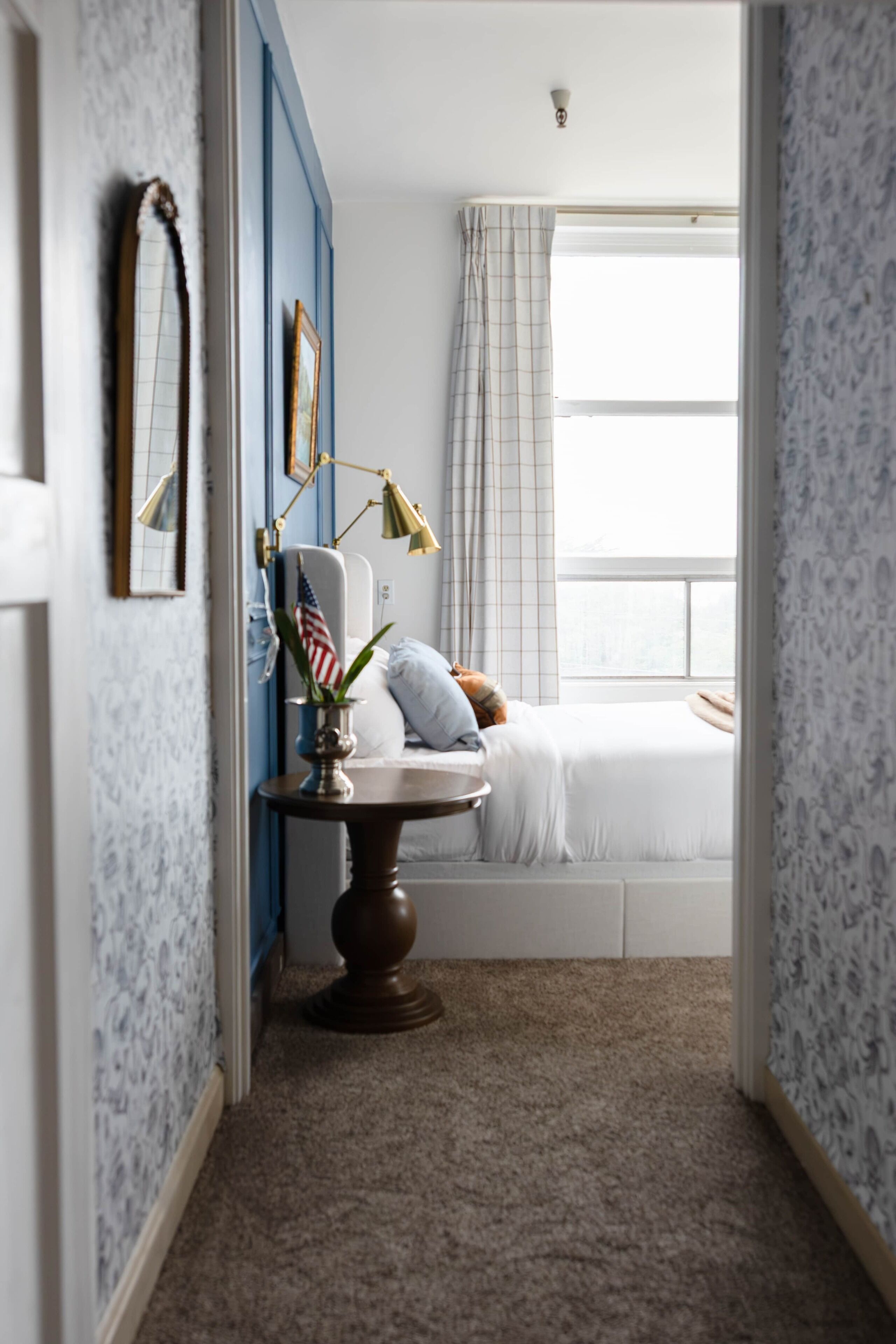Une chambre à coucher avec un lit, une table de chevet avec une lampe, une fenêtre avec des rideaux et un mur orné d’un miroir et de cadres de photos.