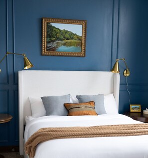 A bedroom with a white bed, blue walls, and a framed landscape painting above the bed.