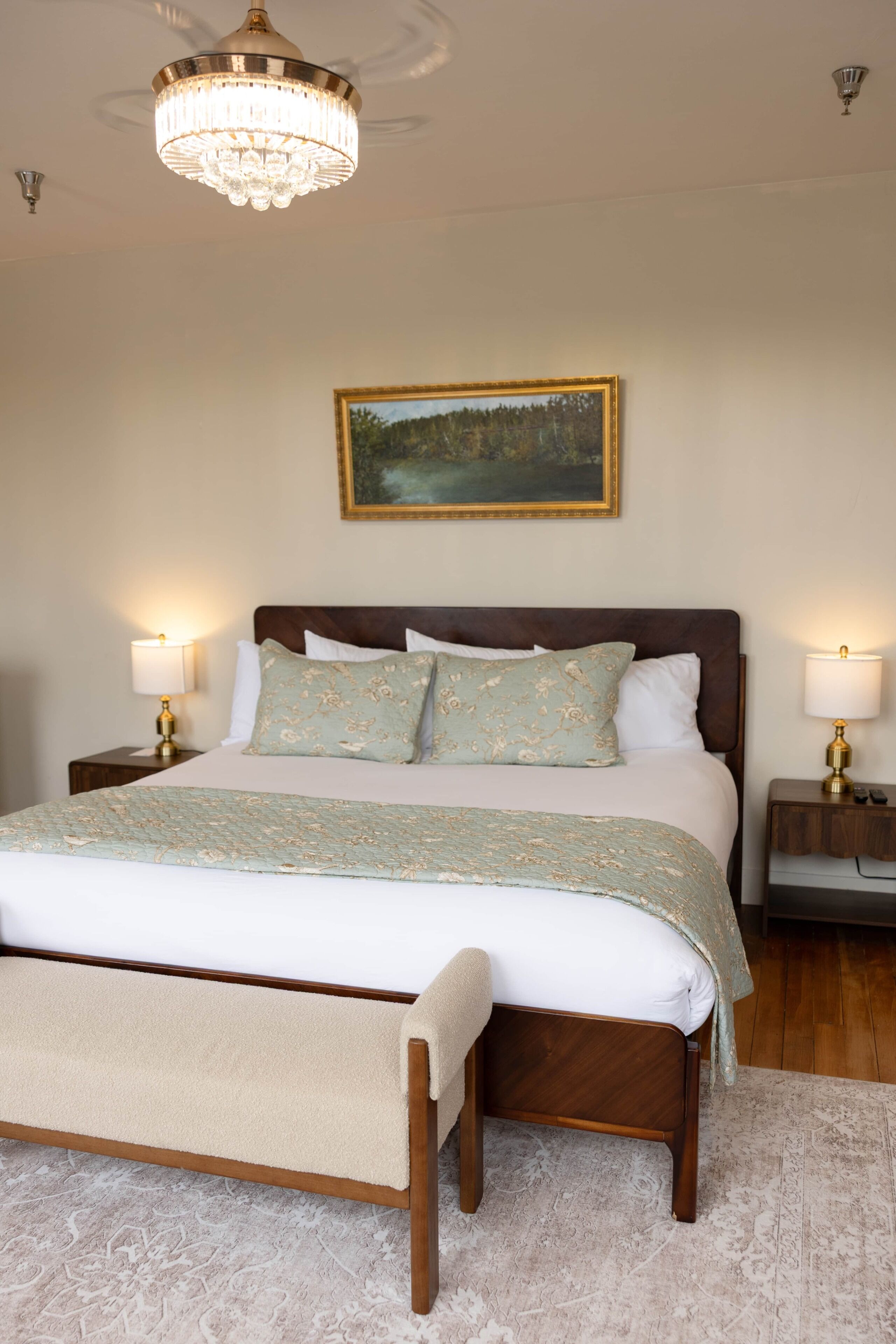 Une chambre à coucher bien aménagée, avec un grand lit, deux tables de chevet avec des lampes, un lustre et un tableau représentant un paysage, accroché au mur.